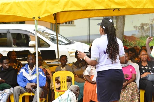 A facilitator stands to address community members seated under a tent.