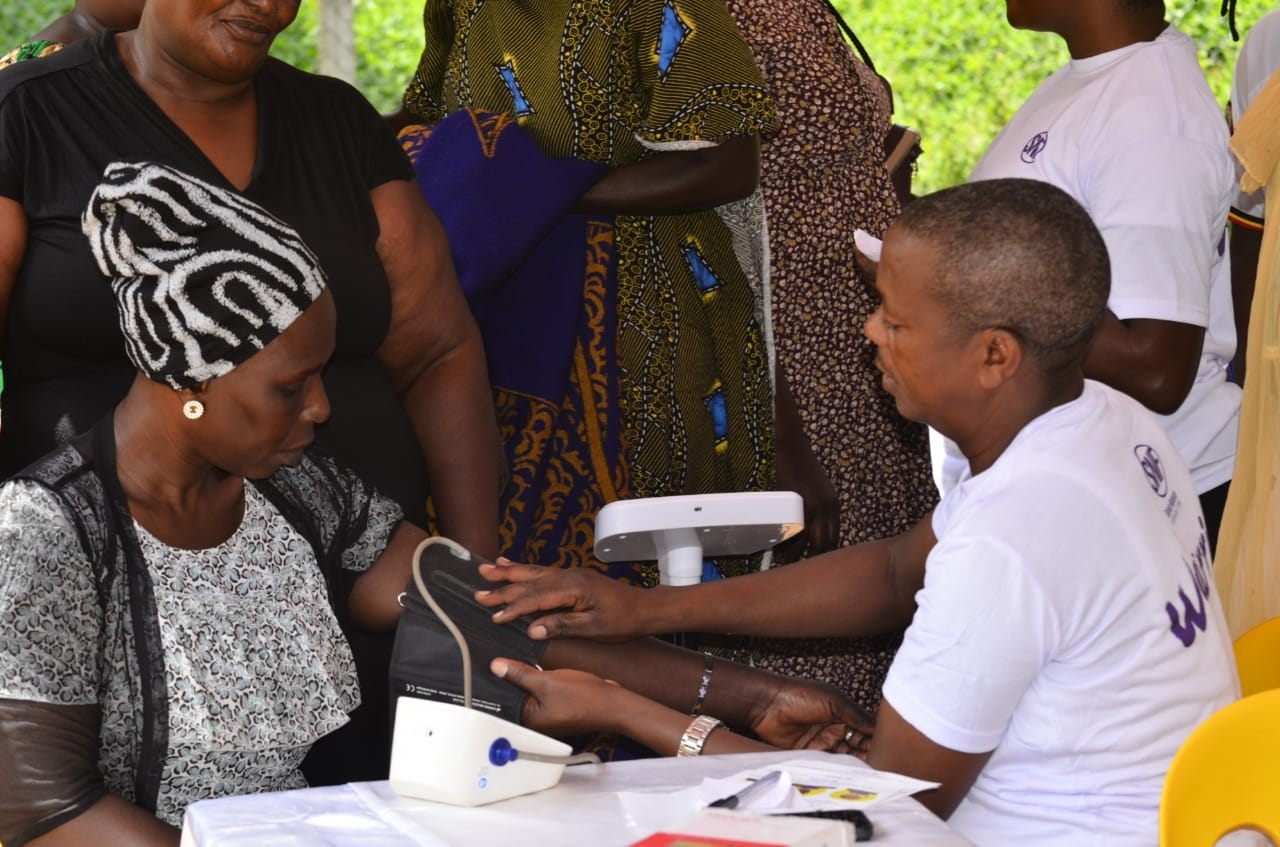 A health worker fits a blood pressure cuff to a woman's arm at an RDAF community screening outreach, while others wait their turn.