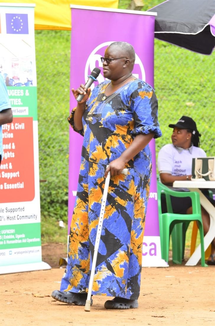 A woman speaks into a microphone at a public health event, supported by a walking cane, with partner banners behind her.